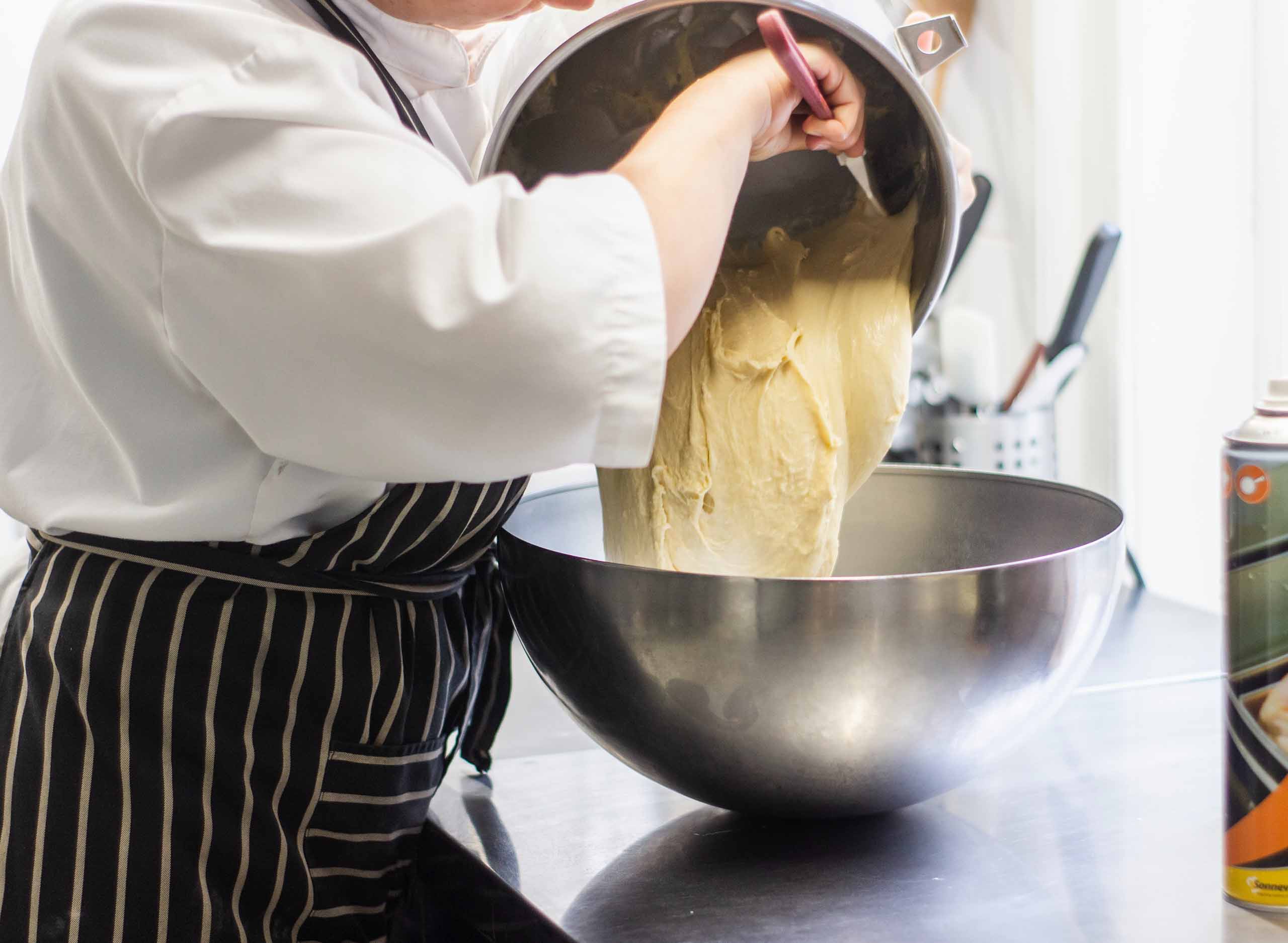 A pastry chef transferring a large amount of dough to a shiny stainless steel bowl.