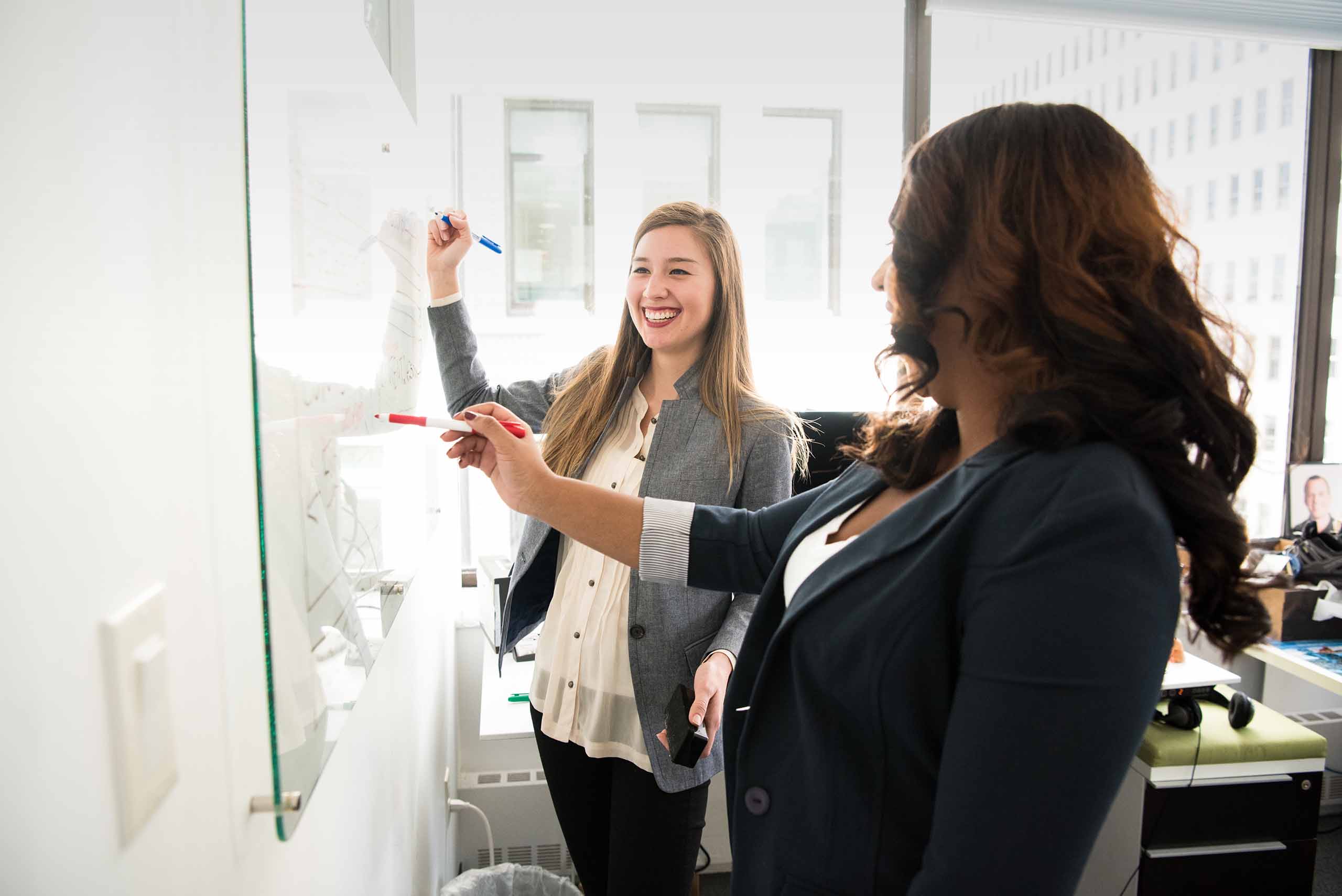 Two women writing on a dry erase whiteboard in an office setting.